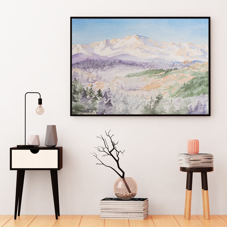 Op de afbeelding: Een ingelijst aquarel schilderij van een besneeuwde bergketen met een bos op de voorgrond. Het schilderij hangt aan een witte muur boven een zwart nachtkastje met een lamp en twee vazen. Een vaas met een tak en een stapel boeken staat op de vloer voor het nachtkastje. Een kleine tafel met een stapel boeken en een roze kaars staat aan de rechterkant van de afbeelding.