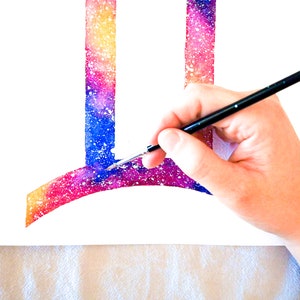 Puede incluir: Una mano sosteniendo un pincel, pintando un dise&ntilde;o de galaxia de acuarela en un lienzo blanco. El dise&ntilde;o es una letra estilizada "I" con una parte superior curva, y los colores son rosa, azul y amarillo.