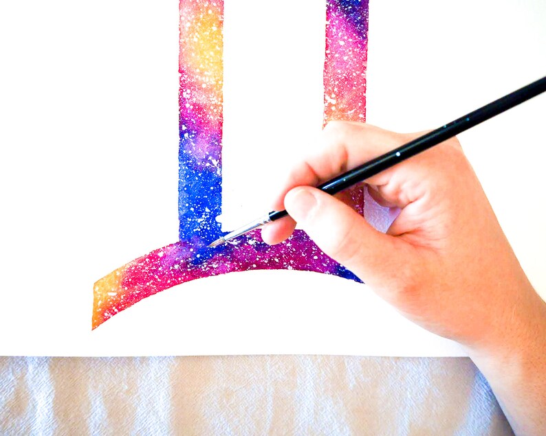 Puede incluir: Una mano sosteniendo un pincel, pintando un dise&ntilde;o de galaxia de acuarela en un lienzo blanco. El dise&ntilde;o es una letra estilizada "I" con una parte superior curva, y los colores son rosa, azul y amarillo.