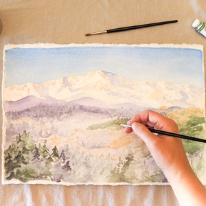 Op de afbeelding: Een aquarel schilderij van een bergketen met besneeuwde toppen. De kunstenaar gebruikt een penseel om details toe te voegen aan de voorgrond, die een bos met bomen laat zien.