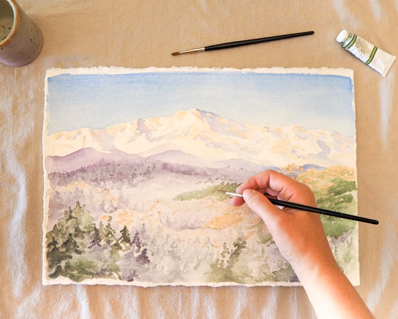 Op de afbeelding: Een aquarel schilderij van een bergketen met besneeuwde toppen. De kunstenaar gebruikt een penseel om details toe te voegen aan de voorgrond, die een bos met bomen laat zien.