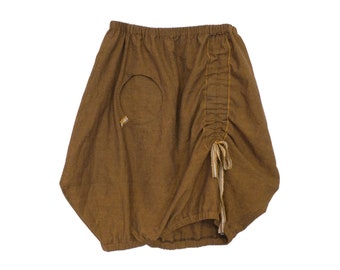 May include: A brown linen skirt with a gathered detail on one side and a circular cut-out pocket on the other side. The skirt has a drawstring tie at the bottom.