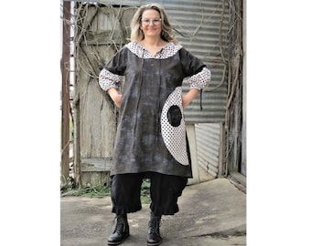 Puede incluir: Una mujer lleva un vestido de lunares negros y blancos con un gran círculo negro en la parte delantera. El vestido tiene un escote fruncido y mangas largas. Está de pie frente a una valla de madera con enredaderas que crecen en ella. El texto "Dottie" está por encima de la imagen y "Opal Annie" está por debajo de la imagen.