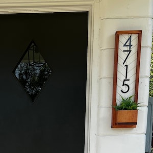 May include: A black front door with a diamond-shaped window is the focal point. A welcome mat with the word "hello" in black script sits in front of the door. A wooden address sign with the numbers "4715" and a small planter box is mounted to the right.