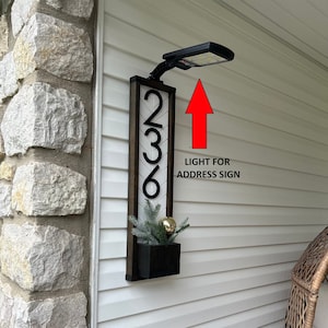 May include: A black and white wooden sign with the numbers 236 on it. The sign is mounted on a white wall with a black light fixture above it. The light fixture is pointing towards the sign. The text "LIGHT FOR ADDRESS SIGN" is written below the light fixture.