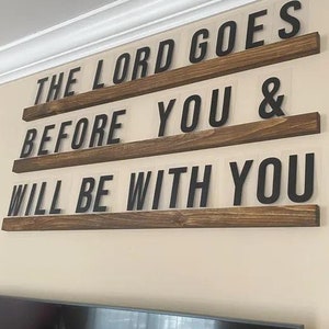 May include: A wooden wall sign with the text "THE LORD GOES BEFORE YOU & WILL BE WITH YOU" in black letters. The letters are mounted on three horizontal, stained wooden shelves against a cream-colored wall. A television is visible below.