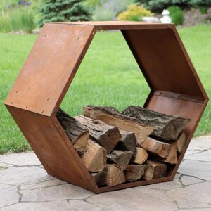 Puede incluir: Un le&ntilde;ero hexagonal de metal marr&oacute;n oxidado lleno de troncos apilados. El dise&ntilde;o de tapa abierta permite un f&aacute;cil acceso a la le&ntilde;a. El soporte se encuentra sobre un patio de piedra con un c&eacute;sped verde al fondo.