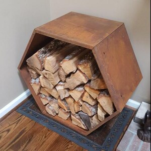 Puede incluir: Un le&ntilde;ero hexagonal de metal marr&oacute;n oxidado lleno de troncos apilados. El soporte se encuentra sobre una alfombra negra decorativa en un piso de madera noble, cerca de una chimenea de ladrillo.