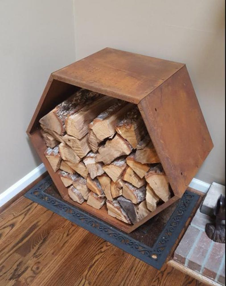 Puede incluir: Un le&ntilde;ero hexagonal de metal marr&oacute;n oxidado lleno de troncos apilados. El soporte se encuentra sobre una alfombra negra decorativa en un piso de madera noble, cerca de una chimenea de ladrillo.