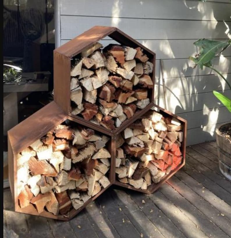 Puede incluir: Un le&ntilde;ero hexagonal de metal marr&oacute;n oscuro. El soporte est&aacute; lleno de le&ntilde;a cortada, mostrando tonos de madera claros y oscuros. El dise&ntilde;o es moderno y funcional, perfecto para el almacenamiento al aire libre.