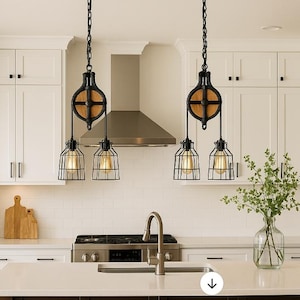 May include: Two black pendant lights with a cage design and Edison bulbs hang from the ceiling. The lights are suspended by a pulley system with a wooden wheel. The kitchen features white cabinets and a stainless steel extractor hood.