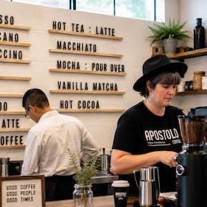 May include: A coffee shop interior featuring a wall-mounted menu board with black letters spelling Espresso, Americano, Cappuccino, Latte, Mocha, Cortado, Chai Latte, Drip Coffee, Hot Tea, Macchiato, Pour Over, Vanilla Matcha, and Hot Cocoa.