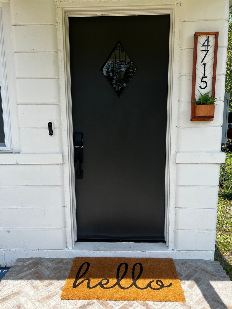 May include: A black front door with a diamond-shaped window is the focal point. A welcome mat with the word "hello" in black script sits in front of the door. A wooden address sign with the numbers "4715" and a small planter box is mounted to the right.