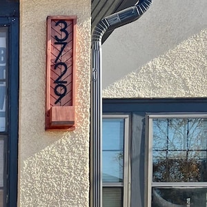 May include: A wooden address sign with the numbers 3729 in black, mounted on a textured wall. The sign has a rectangular shape with a small planter box at the bottom. The wood has a reddish-brown hue.