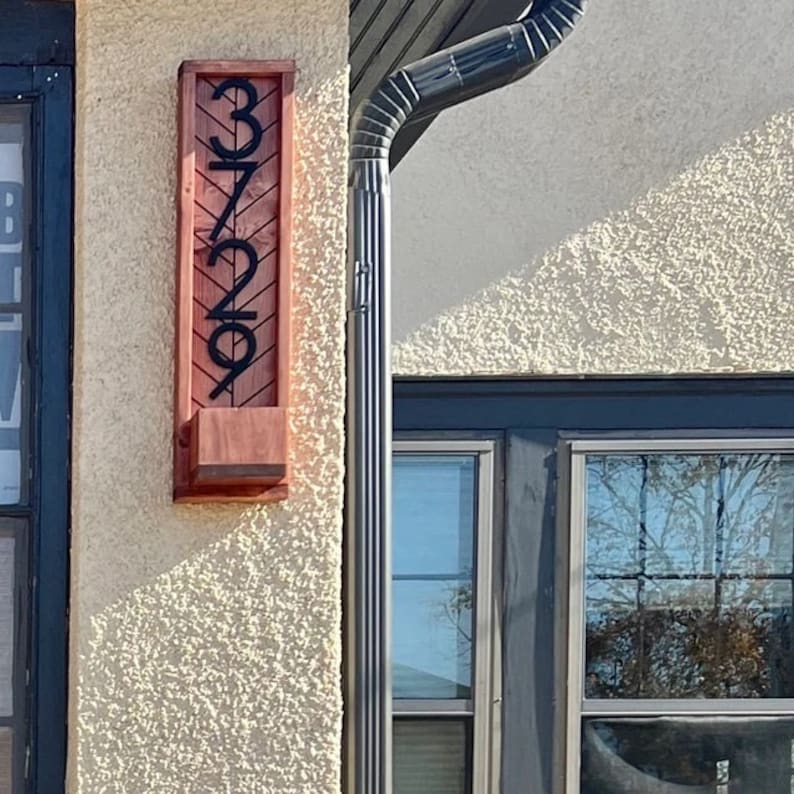 May include: A wooden address sign with the numbers 3729 in black, mounted on a textured wall. The sign has a rectangular shape with a small planter box at the bottom. The wood has a reddish-brown hue.