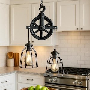 May include: Black, industrial-style pendant lights with a pulley system. The lights feature a cage-like design around the bulbs, suspended from a chain. The kitchen setting includes white cabinets, a stainless steel stove, and a wooden bowl of green apples.