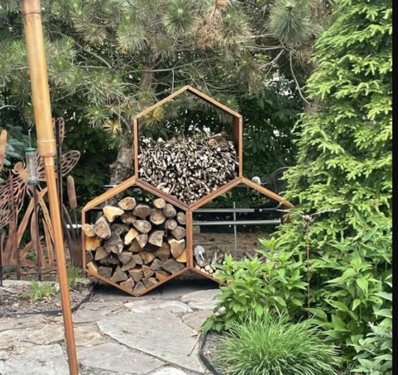 Puede incluir: Una unidad de almacenamiento de le&ntilde;a hexagonal de madera llena de troncos cortados y le&ntilde;a. La unidad est&aacute; al aire libre, rodeada de vegetaci&oacute;n, y se encuentra en un camino de piedra. La madera es de color marr&oacute;n natural.