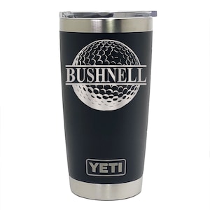 May include: Black Yeti tumbler with a silver lid and a silver golf ball design with the word "BUSHNELL" engraved in the middle. The word "YETI" is engraved on the bottom of the tumbler.