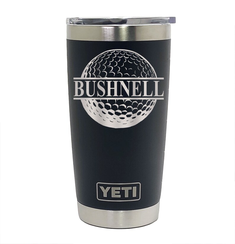 May include: Black Yeti tumbler with a silver lid and a silver golf ball design with the word "BUSHNELL" engraved in the middle. The word "YETI" is engraved on the bottom of the tumbler.