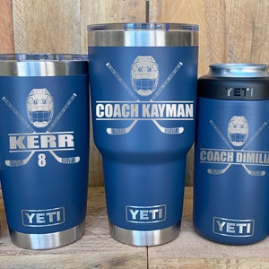 May include: Five blue Yeti tumblers with hockey-themed designs. The tumblers feature a hockey helmet with crossed hockey sticks and personalized text. The text on the tumblers reads "Coach Kayman", "Kerr 8", "Coach Dimilia", and "Coach Pete".