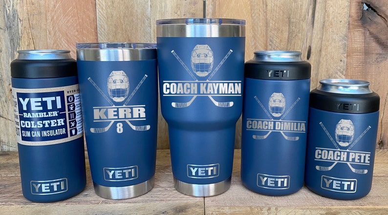 May include: Five blue Yeti tumblers with hockey-themed designs. The tumblers feature a hockey helmet with crossed hockey sticks and personalized text. The text on the tumblers reads "Coach Kayman", "Kerr 8", "Coach Dimilia", and "Coach Pete".