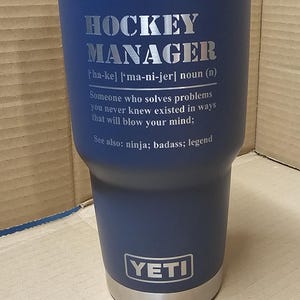 May include: A blue YETI tumbler with a stainless steel lid and a silver inscription that reads "HOCKEY MANAGER [ha-ke] ['ma-ni-jer] noun (n) Someone who solves problems you never knew existed in ways that will blow your mind: See also: ninja; badass; legend"