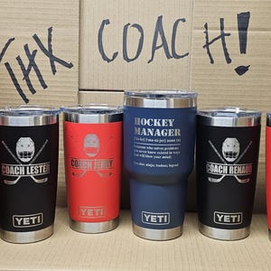May include: Seven stainless steel tumblers with various hockey-themed engravings. The tumblers are in red, black, and blue. The text on the tumblers reads "Coach Today", "Coach Lester", "Coach Terry", "Hockey Manager", "Coach Renaud", and "Coach Smith". The tumblers are arranged in a row on a brown surface. The text "THX COACH!" is written on the surface behind the tumblers.