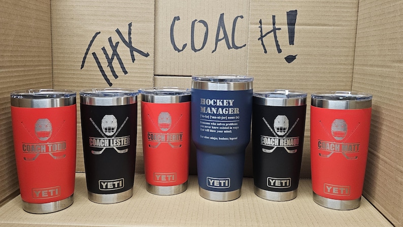 May include: Seven stainless steel tumblers with various hockey-themed engravings. The tumblers are in red, black, and blue. The text on the tumblers reads "Coach Today", "Coach Lester", "Coach Terry", "Hockey Manager", "Coach Renaud", and "Coach Smith". The tumblers are arranged in a row on a brown surface. The text "THX COACH!" is written on the surface behind the tumblers.