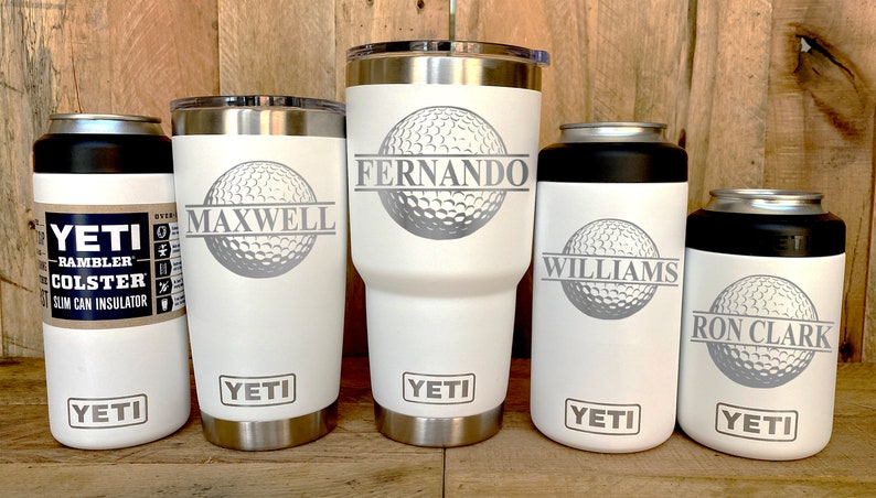 May include: Five white Yeti tumblers with a golf ball design and personalized names. The tumblers are in different sizes, with the largest one being a 30 oz Rambler. The names on the tumblers are Fernando, Maxwell, Williams, and Ron Clark.
