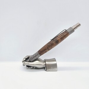 May include: A silver pen with a wood grain barrel and a silver clip. The pen is resting in a silver hand-shaped pen holder.