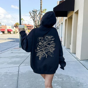 May include: A black hooded sweatshirt with a graphic print of a woman's face with snakes on the back. The sweatshirt is being worn by a person walking down a pavement.