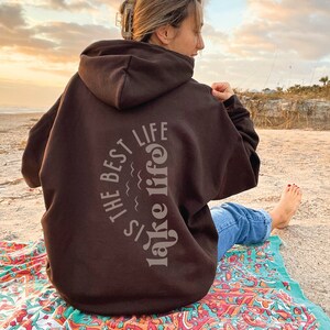 Puede incluir: Una sudadera con capucha marr&oacute;n oscuro con el texto "The Best Lake Life" en un dise&ntilde;o arqueado gris claro en la espalda. La sudadera con capucha la lleva una persona sentada en una toalla de playa colorida en una playa de arena. El cielo est&aacute; nublado.