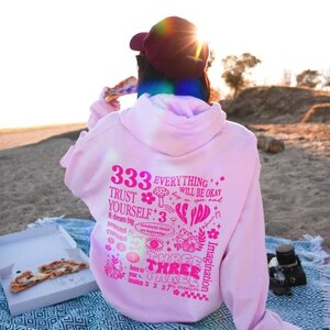 May include: Pink hoodie with a graphic print of various illustrations and text, including "333", "Everything will be okay", "Trust yourself", and "Three Three Three".