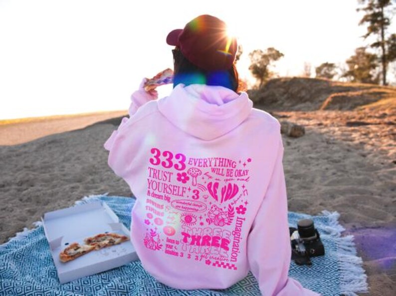 May include: Pink hoodie with a graphic print of various illustrations and text, including "333", "Everything will be okay", "Trust yourself", and "Three Three Three".