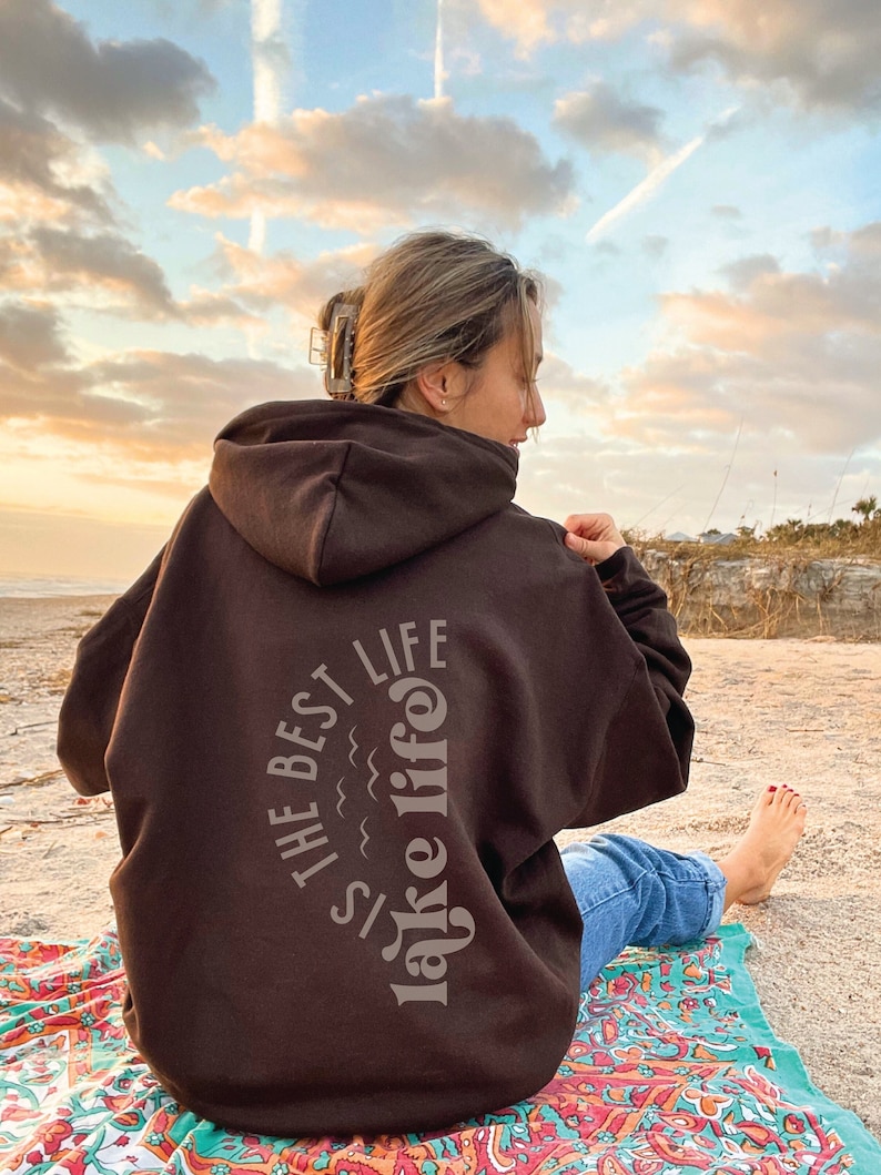 Puede incluir: Una sudadera con capucha marr&oacute;n oscuro con el texto "The Best Lake Life" en un dise&ntilde;o arqueado gris claro en la espalda. La sudadera con capucha la lleva una persona sentada en una toalla de playa colorida en una playa de arena. El cielo est&aacute; nublado.
