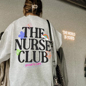 May include: A light grey sweatshirt with the text "THE NURSE CLUB" in black, with colourful graphics. Below the text is "proud to care" in a purple font. A black purse with a silver chain is visible.
