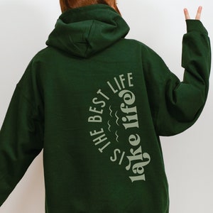 Puede incluir: Sudadera con capucha verde oscuro con el texto "THE BEST LIFE is lake life" en color verde claro. La sudadera tiene capucha y mangas largas. La imagen est&aacute; tomada desde atr&aacute;s, mostrando el dise&ntilde;o del texto en la parte posterior de la sudadera.