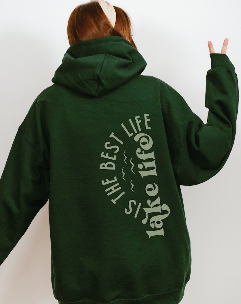 Puede incluir: Sudadera con capucha verde oscuro con el texto "THE BEST LIFE is lake life" en color verde claro. La sudadera tiene capucha y mangas largas. La imagen est&aacute; tomada desde atr&aacute;s, mostrando el dise&ntilde;o del texto en la parte posterior de la sudadera.