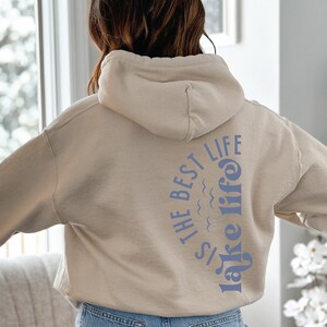 Puede incluir: Una sudadera con capucha beige con el texto "IS THE BEST LIFE lake life" impreso en azul en la espalda. La sudadera con capucha tiene una capucha y mangas largas. La persona lleva vaqueros azules.