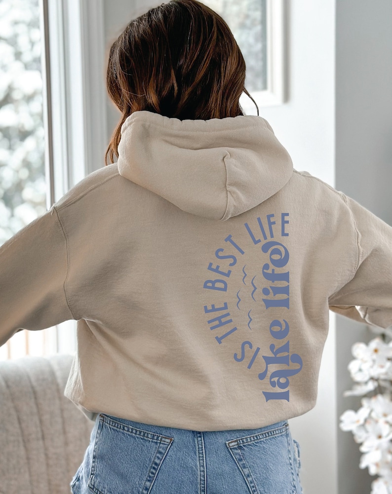 Puede incluir: Una sudadera con capucha beige con el texto "IS THE BEST LIFE lake life" impreso en azul en la espalda. La sudadera con capucha tiene una capucha y mangas largas. La persona lleva vaqueros azules.