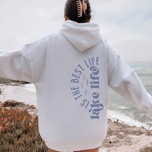 Puede incluir: Una sudadera con capucha blanca con el texto azul "Is the best lake life". La persona lleva una pinza para el pelo. La persona est&aacute; de pie cerca del mar. El texto est&aacute; en un dise&ntilde;o curvo.