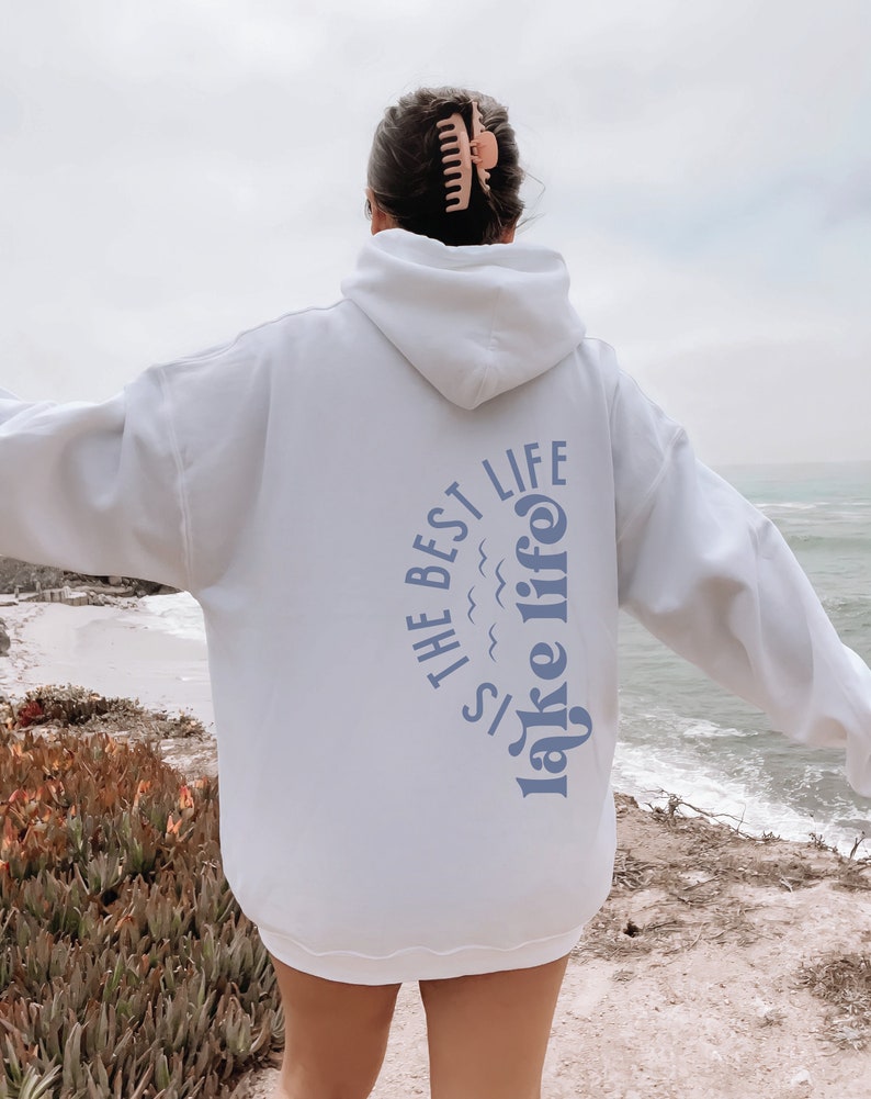 Puede incluir: Una sudadera con capucha blanca con el texto azul "Is the best lake life". La persona lleva una pinza para el pelo. La persona est&aacute; de pie cerca del mar. El texto est&aacute; en un dise&ntilde;o curvo.