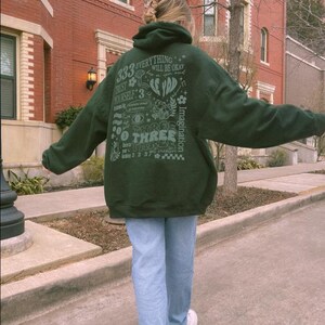 May include: A dark green hoodie with a graphic print on the back. The print features the text "333 Everything Will Be Okay" and other designs. The person is wearing blue jeans and white sneakers.