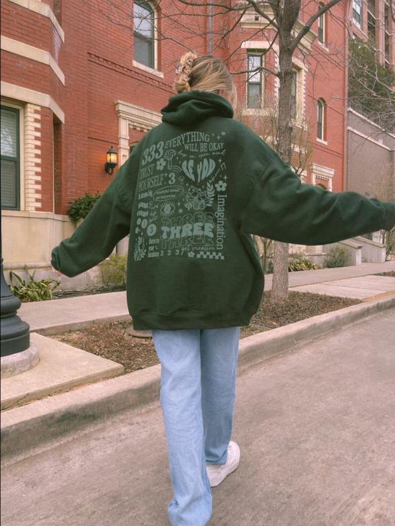 May include: A dark green hoodie with a graphic print on the back. The print features the text "333 Everything Will Be Okay" and other designs. The person is wearing blue jeans and white sneakers.