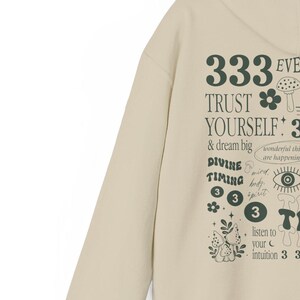 May include: A beige hoodie with a green graphic print featuring the text "333 Trust Yourself 3 & dream big", "Divine Timing 333", and "listen to your intuition 333".
