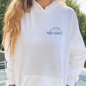 Puede incluir: Sudadera con capucha blanca con bolsillo delantero y capucha. El texto "lake life is the best life" est&aacute; impreso en azul en el pecho. La sudadera con capucha est&aacute; hecha de un material suave.