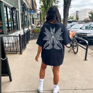 May include: Black oversized t-shirt with a white graphic design of a stylized eye with wings. The graphic is centered on the back of the shirt.