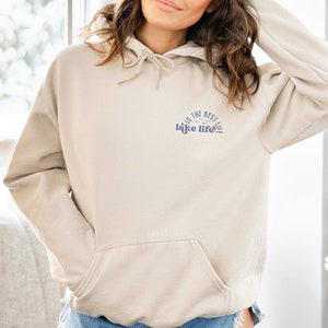 Puede incluir: Sudadera con capucha beige con bolsillo delantero y capucha con cord&oacute;n. En la parte superior izquierda del pecho aparece el texto "this is the best lake life" en azul, con un gr&aacute;fico de olas.