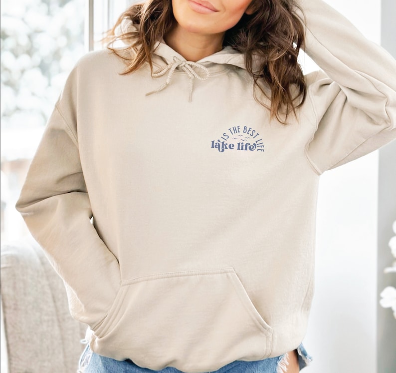 Puede incluir: Sudadera con capucha beige con bolsillo delantero y capucha con cord&oacute;n. En la parte superior izquierda del pecho aparece el texto "this is the best lake life" en azul, con un gr&aacute;fico de olas.