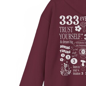 May include: Burgundy hoodie with a white graphic print that says "333 EVE TRUST YOURSELF! & dream big DIVINE TIMING 333 listen to your intuition 3" with illustrations of mushrooms, flowers, and a crescent moon.